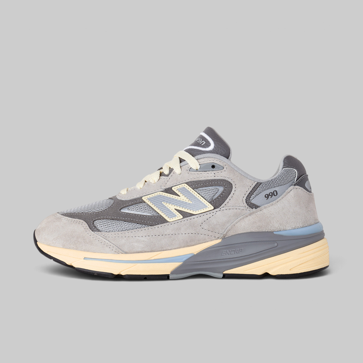 A New Balance 990v3 sneaker in gray suede and mesh with cream and light blue accents, shown in a side profile on a solid gray background.