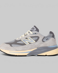 A New Balance 990v3 sneaker in gray suede and mesh with cream and light blue accents, shown in a side profile on a solid gray background.
