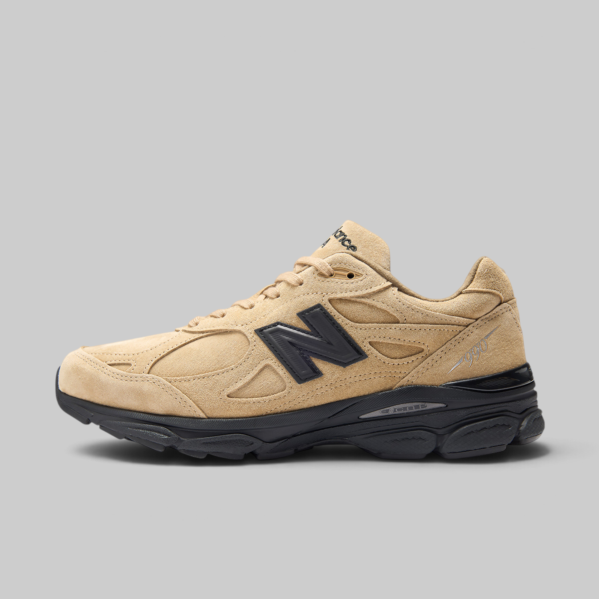 A New Balance sneaker in beige suede and mesh with a prominent black 'N' logo on the side, black laces, and a black midsole, shown against a solid gray background.