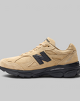 A New Balance sneaker in beige suede and mesh with a prominent black 'N' logo on the side, black laces, and a black midsole, shown against a solid gray background.