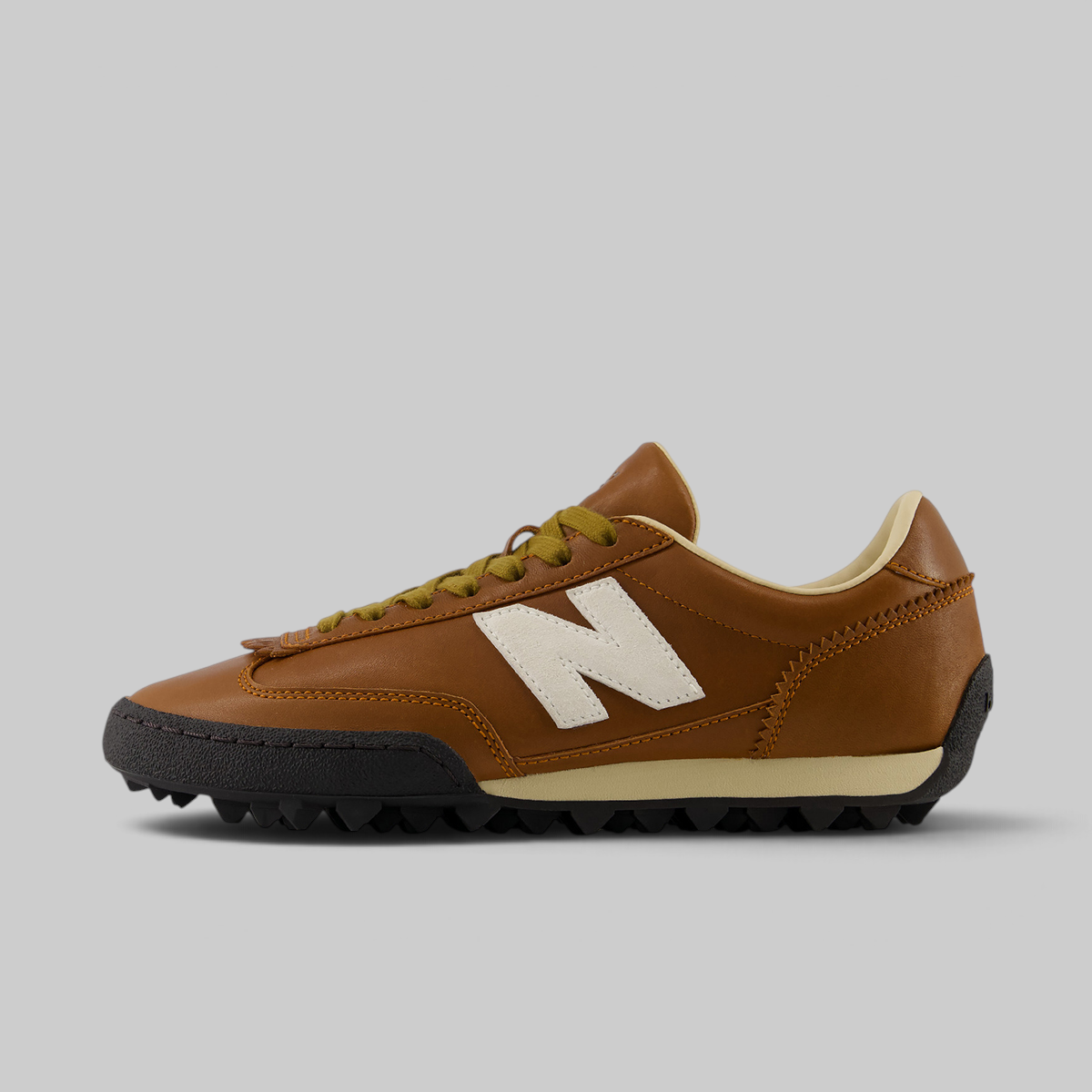 A New Balance low-top sneaker in brown leather with a white 'N' logo, olive green laces, a beige midsole stripe, and a black spiked outsole, shown in a side profile on a light gray background.