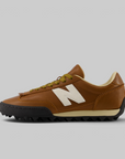 A New Balance low-top sneaker in brown leather with a white 'N' logo, olive green laces, a beige midsole stripe, and a black spiked outsole, shown in a side profile on a light gray background.