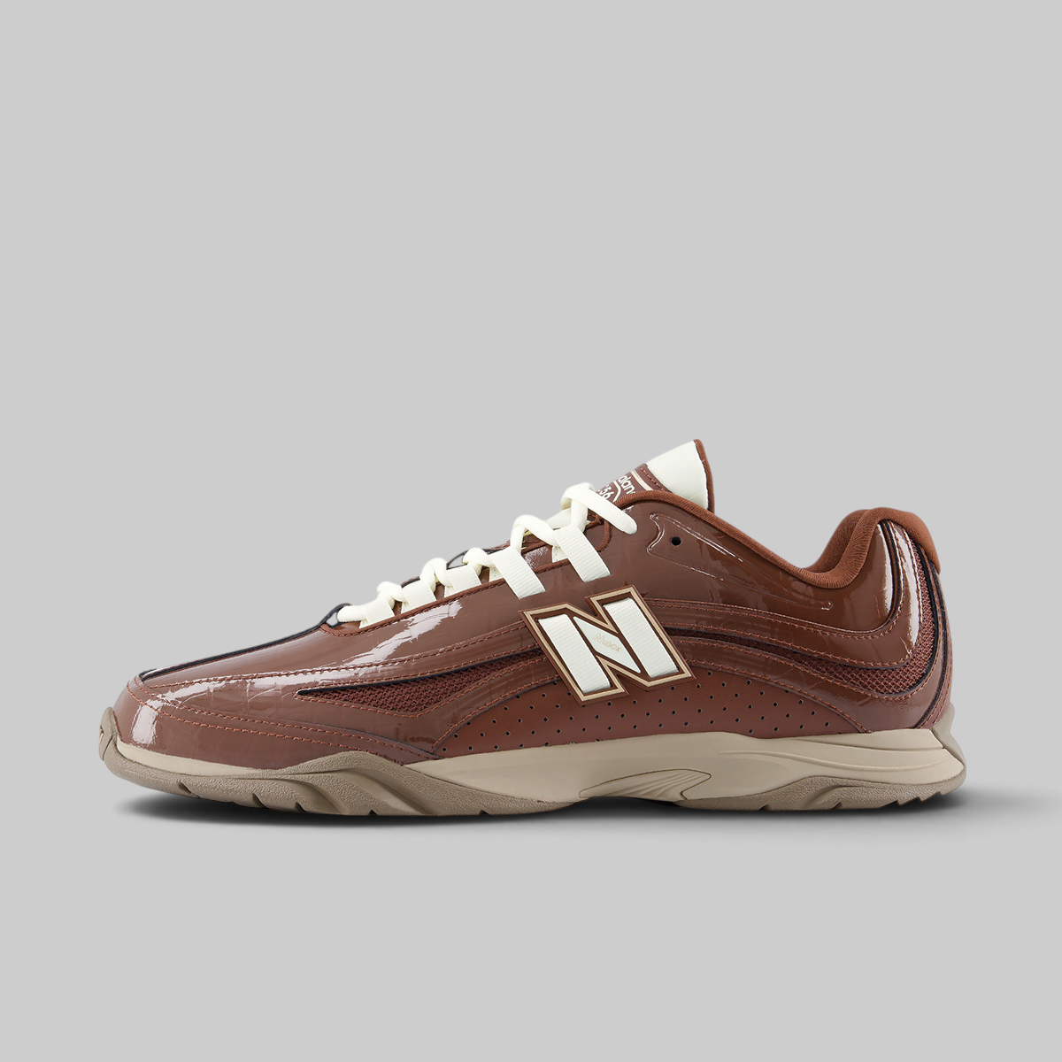 A New Balance low-top sneaker in brown patent leather and mesh, featuring white laces and a large white 'N' logo with a gold outline on the side, set against a solid gray background.