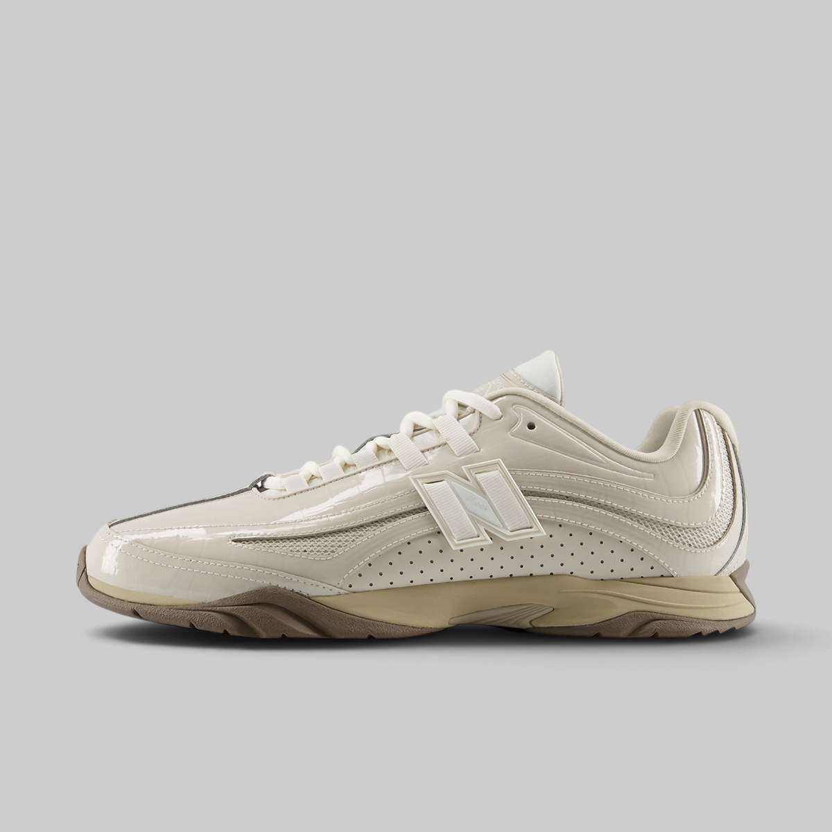 A New Balance athletic shoe in cream and beige with a glossy patent leather upper, perforated side panels, and a chunky beige midsole, shown in a side profile on a solid gray background.