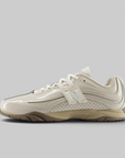 A New Balance athletic shoe in cream and beige with a glossy patent leather upper, perforated side panels, and a chunky beige midsole, shown in a side profile on a solid gray background.